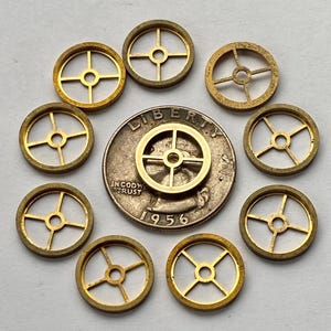 May include: A collection of small, gold-colored watch balance wheels arranged around a silver coin. The coin is dated 1956 and features the words "Liberty" and "In God We Trust". The balance wheels have a circular design with four spokes.