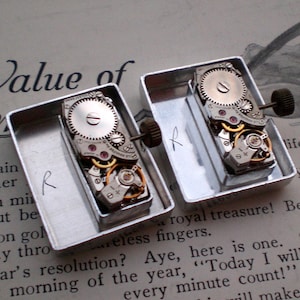 May include: Two vintage watch movements displayed in small, open metal containers. The intricate mechanisms feature silver gears, gold accents, and red jewels. The background includes text from a vintage document.