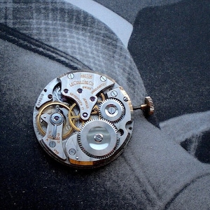 Pu&ograve; includere: Primo piano di un movimento di orologio vintage con ingranaggi e componenti intricati. Il meccanismo dell'orologio presenta il testo "LeCoultre & Co. Swiss Seventeen 17 Jewels Unadjusted". Le parti metalliche sono argentate e dorate.