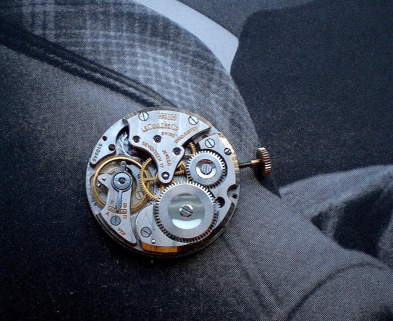 Pu&ograve; includere: Primo piano di un movimento di orologio vintage con ingranaggi e componenti intricati. Il meccanismo dell'orologio presenta il testo "LeCoultre & Co. Swiss Seventeen 17 Jewels Unadjusted". Le parti metalliche sono argentate e dorate.