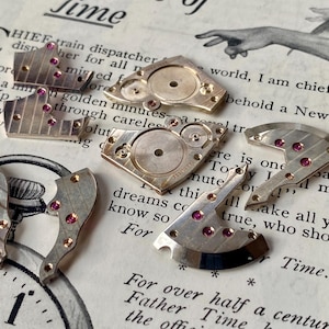 May include: Close-up of disassembled watch parts. Silver-toned metal components with small red jewels are scattered on a vintage paper background with printed text and a clock face.