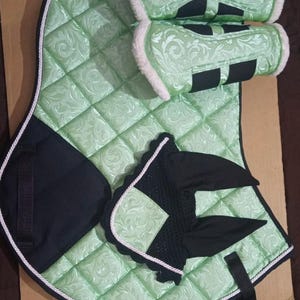 May include: A light green quilted equestrian saddle pad set with matching leg wraps and a fly bonnet. The set features a floral pattern, black accents, and white trim. The leg wraps have a soft, white lining.