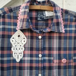 May include: A plaid button-down shirt in shades of blue, pink, and brown, with a white lace applique and pink lace trim on the collar. The shirt has a pocket and a pink button. The tag reads "American Eagle Outfitters".
