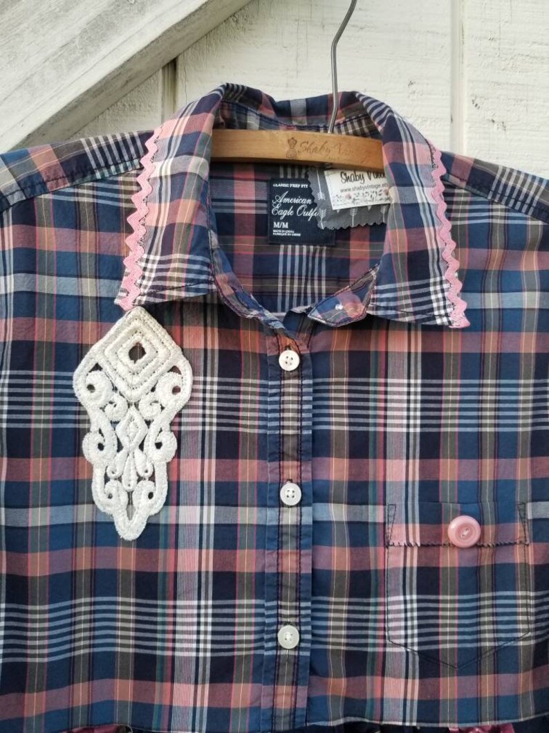May include: A plaid button-down shirt in shades of blue, pink, and brown, with a white lace applique and pink lace trim on the collar. The shirt has a pocket and a pink button. The tag reads "American Eagle Outfitters".