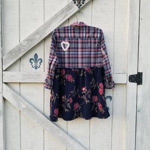 May include: A patchwork dress with a plaid top and a floral print skirt is hanging on a white wooden door. The top is a mix of pink, blue, and white plaid, and the skirt is navy blue with pink flowers. A white heart-shaped patch is sewn on the back.