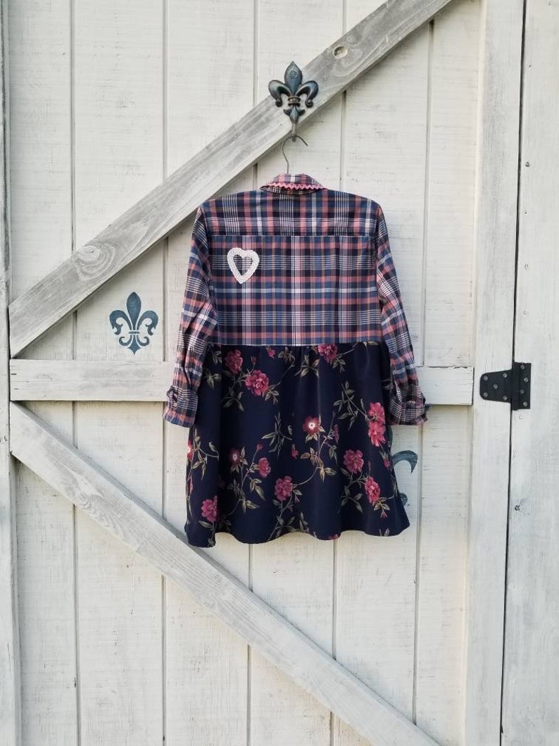 May include: A patchwork dress with a plaid top and a floral print skirt is hanging on a white wooden door. The top is a mix of pink, blue, and white plaid, and the skirt is navy blue with pink flowers. A white heart-shaped patch is sewn on the back.