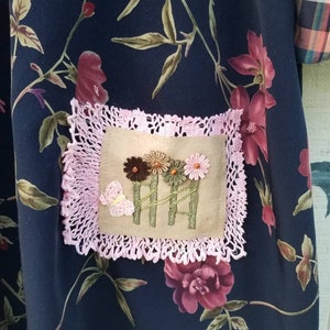 May include: Dark navy fabric with a floral pattern featuring burgundy and pink flowers and green leaves. A square patch with embroidered flowers and a butterfly is framed by a pink lace border. The fabric appears to be part of a garment.