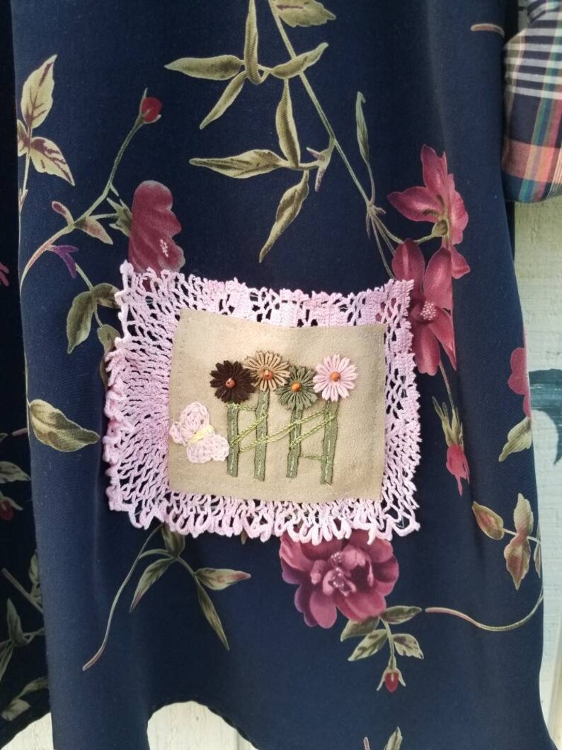 May include: Dark navy fabric with a floral pattern featuring burgundy and pink flowers and green leaves. A square patch with embroidered flowers and a butterfly is framed by a pink lace border. The fabric appears to be part of a garment.
