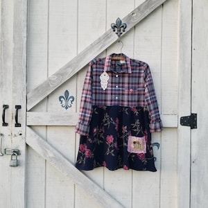 May include: A plaid shirt dress with a navy blue floral skirt. The top half is a pink and navy plaid pattern, and the skirt features pink roses. A decorative patch is sewn on the skirt. The dress is hanging on a wooden hanger.