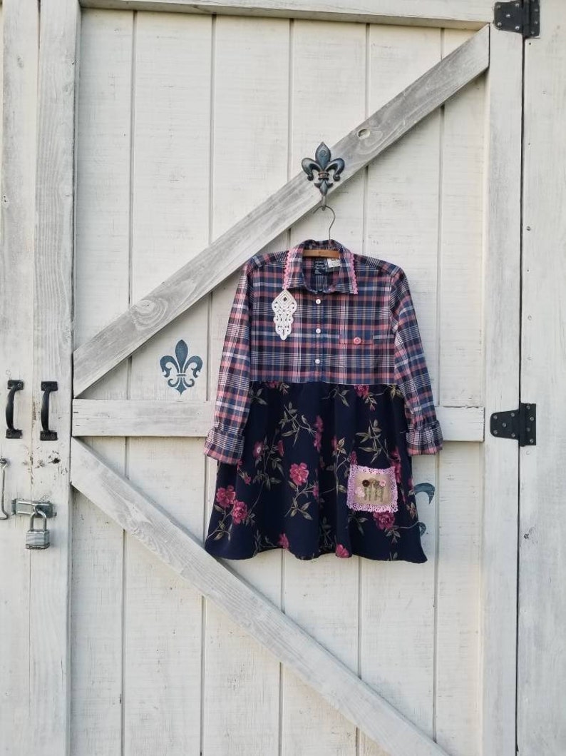 May include: A plaid shirt dress with a navy blue floral skirt. The top half is a pink and navy plaid pattern, and the skirt features pink roses. A decorative patch is sewn on the skirt. The dress is hanging on a wooden hanger.