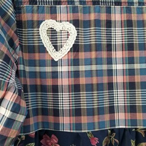May include: A patchwork shirt with a plaid pattern in shades of blue, pink, and white. A white, crocheted heart is sewn onto the back. The bottom of the shirt features a dark blue floral fabric.