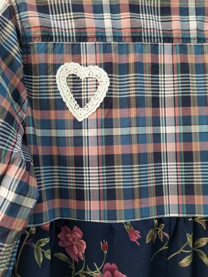 May include: A patchwork shirt with a plaid pattern in shades of blue, pink, and white. A white, crocheted heart is sewn onto the back. The bottom of the shirt features a dark blue floral fabric.
