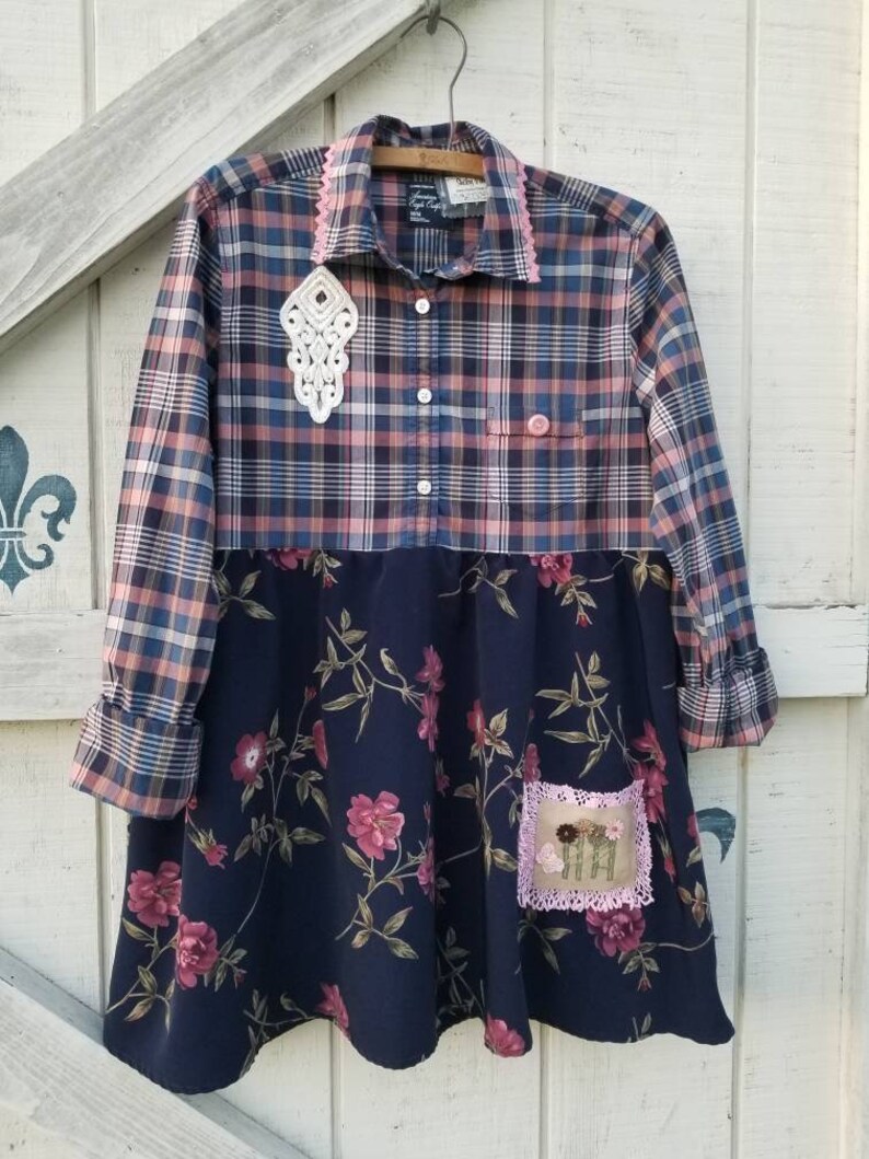 May include: A handmade dress with a plaid top in shades of blue, pink, and beige, and a navy blue floral skirt. The dress features a white lace embellishment and a small embroidered patch with flowers. The collar and pocket are trimmed with pink lace.