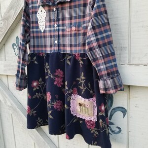 May include: A handmade dress featuring a plaid top in blue, pink, and white, paired with a navy floral skirt. The dress has a pink lace collar and a decorative white lace accent. A small patch with embroidered flowers is sewn on the skirt.