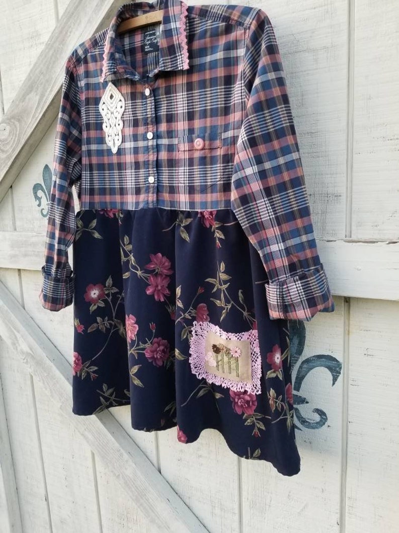 May include: A handmade dress featuring a plaid top in blue, pink, and white, paired with a navy floral skirt. The dress has a pink lace collar and a decorative white lace accent. A small patch with embroidered flowers is sewn on the skirt.