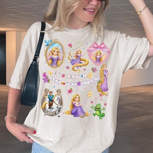May include: A beige short-sleeve t-shirt featuring a graphic print of Rapunzel, Flynn Rider, and Pascal. The design includes various illustrations of the characters, floral accents, and the text "AT LAST I SEE THE LIGHT" in a colorful font.