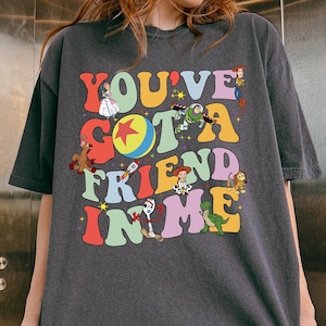 May include: Dark gray t-shirt with the text "YOU'VE GOT A FRIEND IN ME" in colorful, retro-style lettering. The shirt features cartoon characters from a popular animated movie. The shirt is worn by a person.