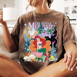 May include: Brown t-shirt featuring a colorful graphic of characters from "The Little Mermaid" movie. The design includes Ariel, Flounder, Sebastian, Ursula, and King Triton. The shirt is worn by a person sitting on a beige couch.