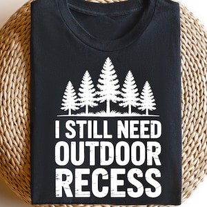 May include: Black t-shirt with a white graphic of pine trees and the text "I STILL NEED OUTDOOR RECESS". The shirt is displayed on a round, woven surface.