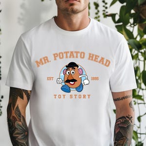 Può includere: T-shirt bianca con grafica di Mr. Potato Head di Toy Story. Il design include il testo "MR. POTATO HEAD" sopra il personaggio, con "EST 1995" e "TOY STORY" sotto. La grafica è nei toni del marrone, blu e nero.
