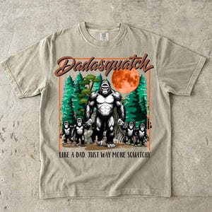 Può includere: Una t-shirt beige a maniche corte con la grafica di un grande gorilla e quattro gorilla più piccoli in una foresta con una luna rossa. Il testo Dadasquatch è in alto e LIKE A DAD, JUST WAY MORE SQUATCHY è in basso in caratteri marroni.