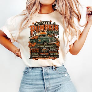 Könnte beinhalten: Ein cremefarbenes Kurzarm-T-Shirt mit einer Grafik eines grünen Oldtimer-Trucks auf einem Kürbisfeld. Der Text lautet Mickey's Pumpkin Patch, Fun for the Whole Family und listet Aktivitäten wie Hayrides, Apple Cider und Memories Grow Here auf.