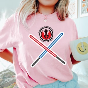 Può includere: Una maglietta rosa chiaro con una grafica di due spade laser incrociate, una rossa e una blu, e un emblema circolare con la scritta "Defenders of the Republic" e "Heroes of the Galaxy". Pantaloncini azzurri.