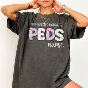 以下が含まれることがあります： 「TINY PATIENTS. BIG HEART'S PEDS nurse」の文字が入った濃いグレーのTシャツ。「PEDS」の文字は花柄とストライプのデザイン。ゆったりとしたフィット感のシャツです。