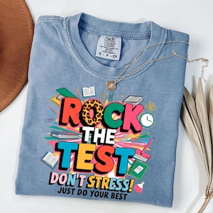 May include: A light blue t-shirt with the words "ROCK THE TEST" in a colorful design. The shirt also says "DON'T STRESS! JUST DO YOUR BEST". A gold necklace is draped across the shirt.