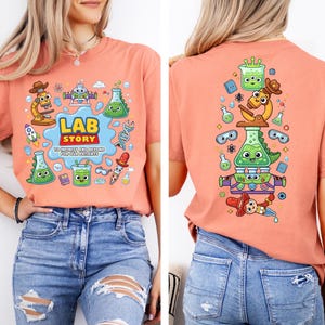 Op de afbeelding: Koraalrood T-shirt met een kleurrijke afbeelding met laboratorium-thema cartoonfiguren en de tekst "LAB STORY". De achterkant van de shirt heeft een bijpassend ontwerp. Het shirt wordt gecombineerd met een blauwe jeans.