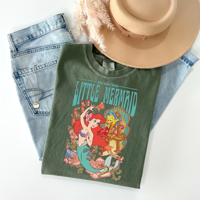 May include: An olive green t-shirt featuring the text "Little Mermaid" with a graphic of Ariel and Flounder. The shirt is paired with light wash denim jeans, a gold necklace, and a tan hat with a brown band. Dried flowers are also present.