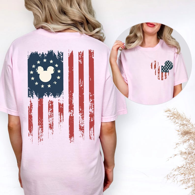 Puede incluir: Una camiseta rosa claro con un gr&aacute;fico de la bandera estadounidense desgastada en la espalda, con una silueta central de cabeza de rat&oacute;n rodeada de estrellas. El pecho izquierdo muestra un gr&aacute;fico m&aacute;s peque&ntilde;o de cabeza de rat&oacute;n con patr&oacute;n de bandera.