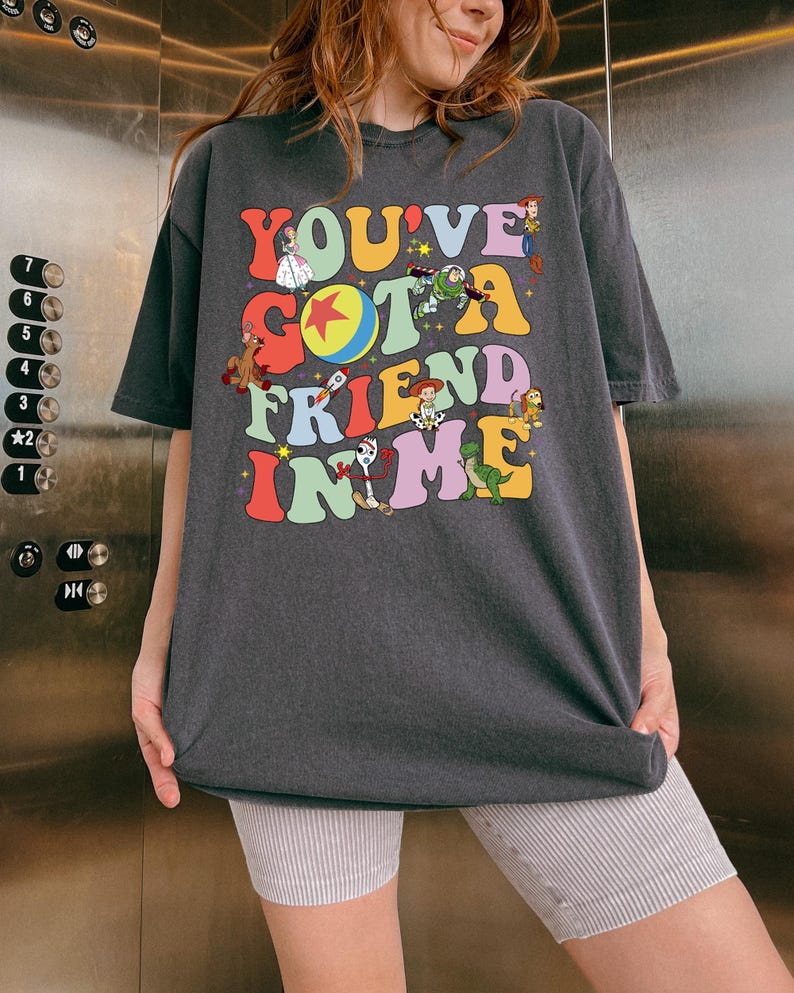 May include: Dark gray t-shirt with the text "YOU'VE GOT A FRIEND IN ME" in colorful, retro-style lettering. The shirt features cartoon characters from a popular animated movie. The shirt is worn by a person.