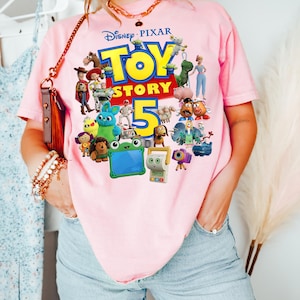 May include: A light pink t-shirt featuring a colorful graphic print of the Disney Pixar Toy Story 5 logo surrounded by various animated characters including Woody, Buzz Lightyear, Bo Peep, Mr. and Mrs. Potato Head, and several other movie figures.