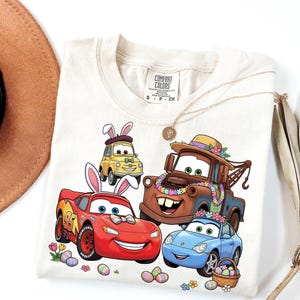 Può includere: T-shirt color crema con auto dei cartoni animati con orecchie da coniglietto pasquale e decorazioni floreali. Le auto sono rosse, gialle, marroni e blu, con uova di Pasqua e fiori. La maglietta ha un'etichetta Comfort Colors.