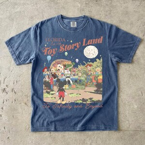 May include: A blue short-sleeve t-shirt featuring a graphic print with the text Florida Est. 2018 Toy Story Land and To Infinity and Beyond. The illustration depicts Mickey Mouse, Woody, Buzz Lightyear, and other characters in a park setting.