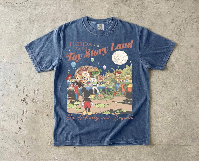 May include: A blue short-sleeve t-shirt featuring a graphic print with the text Florida Est. 2018 Toy Story Land and To Infinity and Beyond. The illustration depicts Mickey Mouse, Woody, Buzz Lightyear, and other characters in a park setting.