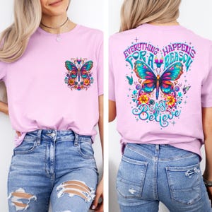 May include: A light pink t-shirt featuring a colorful butterfly design on the front pocket and back. The back of the shirt has the text "Everything Happens For A Reason, Just Believe" in a whimsical font. The butterfly has blue, orange, and yellow accents.