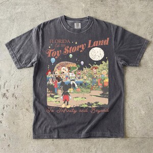 May include: A charcoal gray short-sleeve t-shirt featuring a graphic print with the text Florida Est. 2018 Toy Story Land and To Infinity and Beyond. The illustration depicts characters including Mickey Mouse, Woody, Buzz Lightyear, and Slinky Dog.