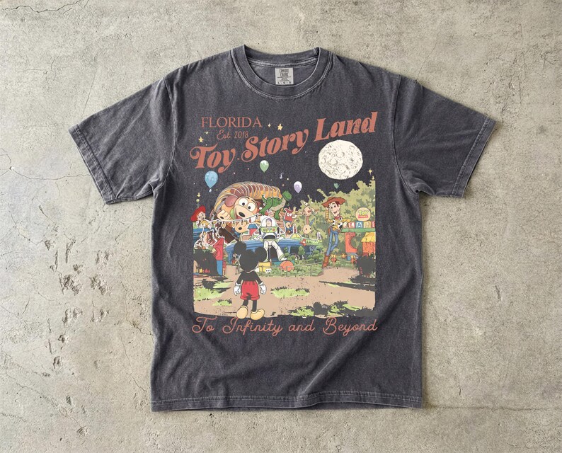 May include: A charcoal gray short-sleeve t-shirt featuring a graphic print with the text Florida Est. 2018 Toy Story Land and To Infinity and Beyond. The illustration depicts characters including Mickey Mouse, Woody, Buzz Lightyear, and Slinky Dog.