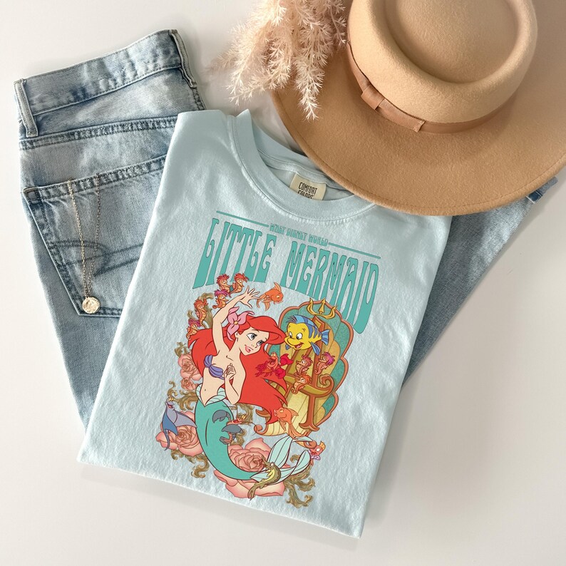 May include: A light blue t-shirt featuring the words "Little Mermaid" in teal lettering, with a graphic of Ariel and Flounder. The shirt is paired with light wash denim jeans and a tan hat.