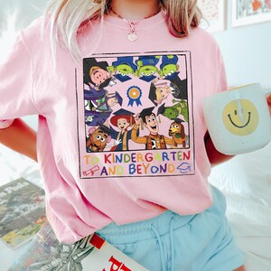 May include: A light pink t-shirt featuring a square graphic of Toy Story characters wearing graduation caps. The text reads TO KINDERGARTEN AND BEYOND with a diploma and graduation cap icon. The characters include Woody, Buzz, and others.
