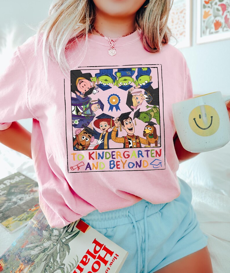 May include: A light pink t-shirt featuring a square graphic of Toy Story characters wearing graduation caps. The text reads TO KINDERGARTEN AND BEYOND with a diploma and graduation cap icon. The characters include Woody, Buzz, and others.