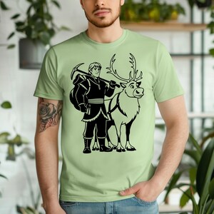 May include: Light green t-shirt featuring a black graphic of a character with a pickaxe and a reindeer. The character is wearing a winter outfit. The reindeer has antlers and a harness.