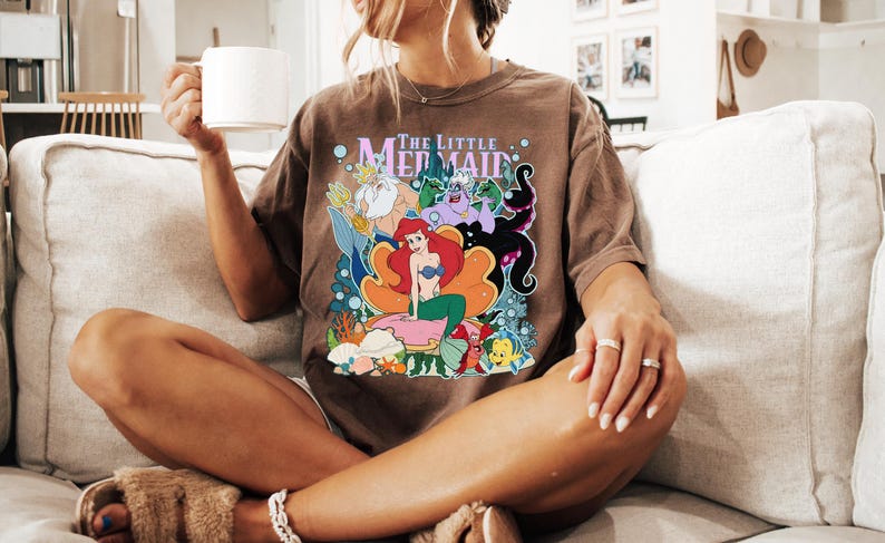 May include: Brown t-shirt featuring a colorful graphic of characters from "The Little Mermaid" movie. The design includes Ariel, Flounder, Sebastian, Ursula, and King Triton. The shirt is worn by a person sitting on a beige couch.