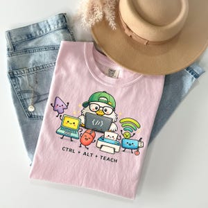Könnte beinhalten: Ein hellrosa T-Shirt mit einer Grafik einer Cartoon-Eule, die eine grüne Kappe und eine Brille trägt, umgeben von animiertem Computerzubehör wie Laptop, Maus, Drucker und WLAN-Symbol, mit dem darunter gedruckten Text CTRL + ALT + TEACH.