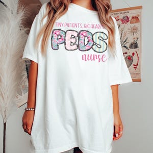 以下が含まれることがあります： 「TINY PATIENTS. BIG HEARTS. PEDS nurse」の文字が、花柄、ギンガムチェック、ストライプ柄でデザインされた白いTシャツ。半袖でゆったりとしたフィット感があり、カジュアルウェアに適しています。