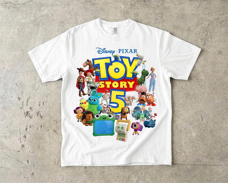 May include: A white short-sleeve t-shirt featuring a colorful graphic print of various characters from the Disney Pixar Toy Story franchise. The central design includes the text Toy Story 5 in large blue and yellow letters surrounded by a group of animated figures.