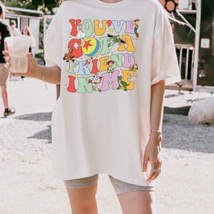 May include: Cream-colored oversized t-shirt featuring the phrase "YOU'VE GOT A FRIEND IN ME" in colorful, retro-style lettering. The design includes cartoon characters. The shirt is a relaxed fit, suitable for casual wear.