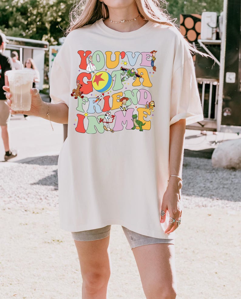 May include: Cream-colored oversized t-shirt featuring the phrase "YOU'VE GOT A FRIEND IN ME" in colorful, retro-style lettering. The design includes cartoon characters. The shirt is a relaxed fit, suitable for casual wear.